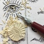 Hand-carved linocut of an eye surrounded by shavings and sketchbook illustrations. A visual metaphor for the creative beginnings of Buen Ojo.